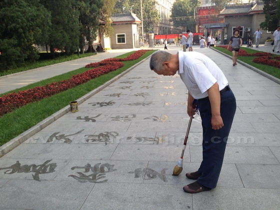 An old man is practicing Chinese caligraphy by writing Chinese characters on the ground with a large brush using water instead of ink